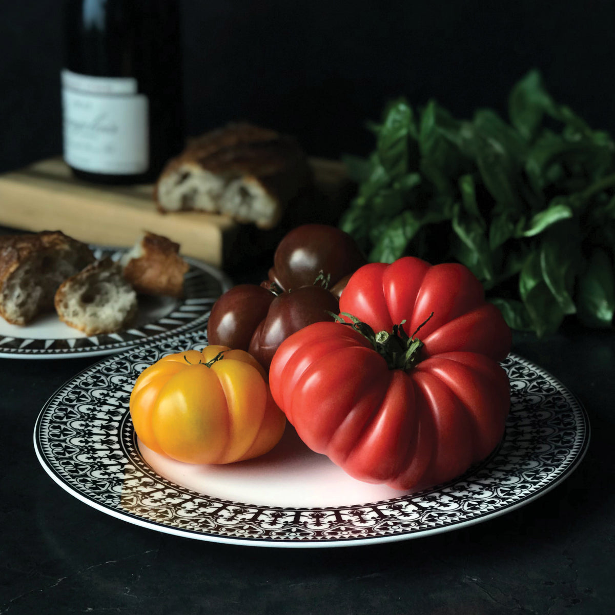 A variety of tomatoes, including a large red one, a yellow one, and brown ones, are arranged on a decorative Casablanca Rimmed Dinner Plate by Caskata Artisanal Home. In the background, there&#39;s a loaf of bread, some basil, and a bottle of wine. The hand decorated details on the high-fired porcelain add an exquisite touch to the scene.