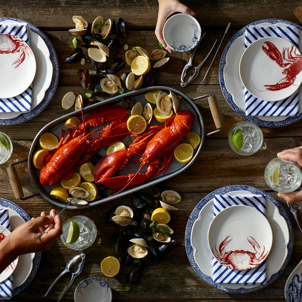 A dining table set with heirloom-quality dinnerware featuring blue and white dishes. Two whole lobsters on a platter with lemon slices are surrounded by mussels, all served on elegant Crab Red Small Plates by Caskata Artisanal Home. Glasses of water with lime complete the scene as four hands reach from the sides.