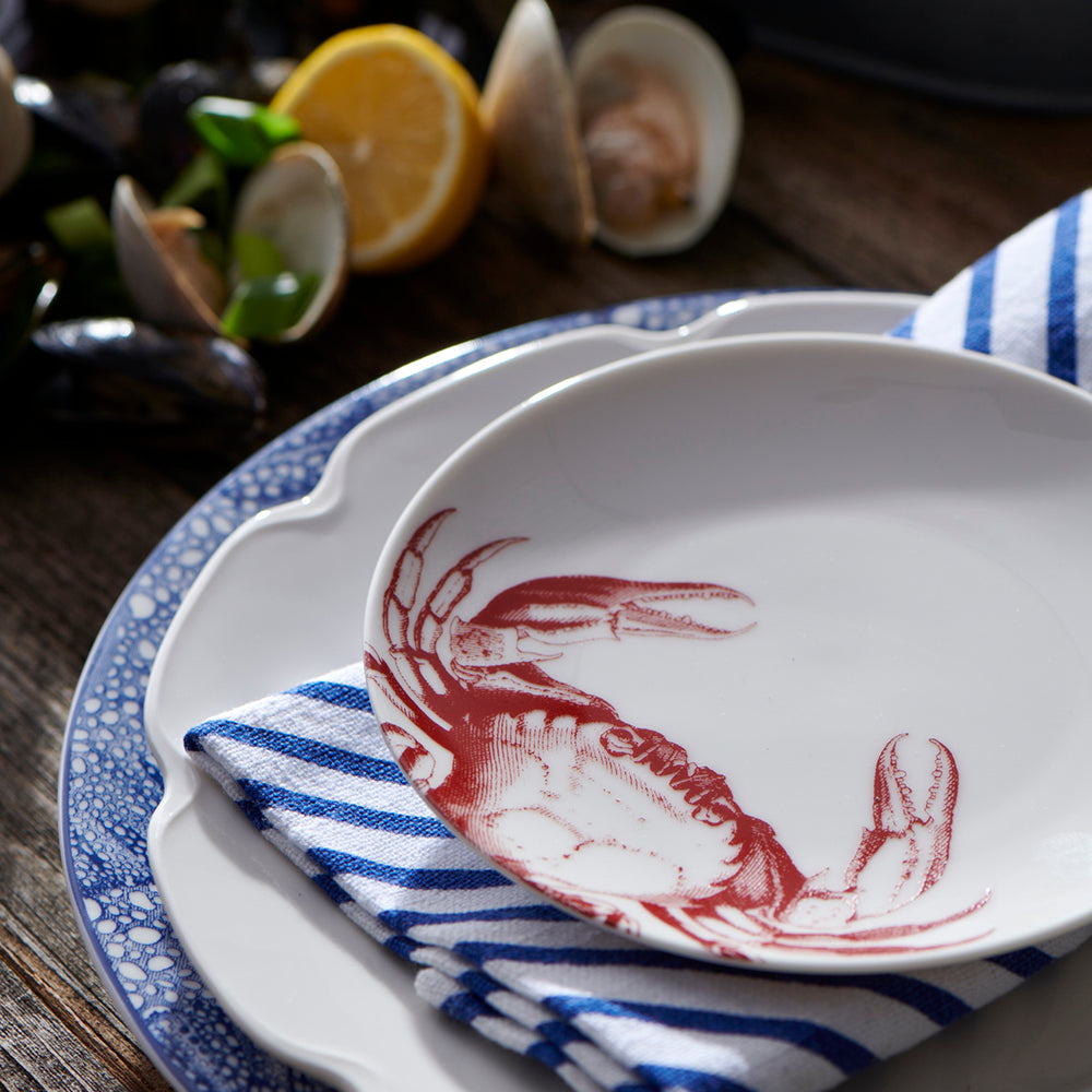 A Caskata Artisanal Home Crab Red Small Plate, part of our heirloom-quality dinnerware collection, sits on a blue and white striped napkin, placed on a larger patterned plate. In the background, there are lemons and open clam shells.