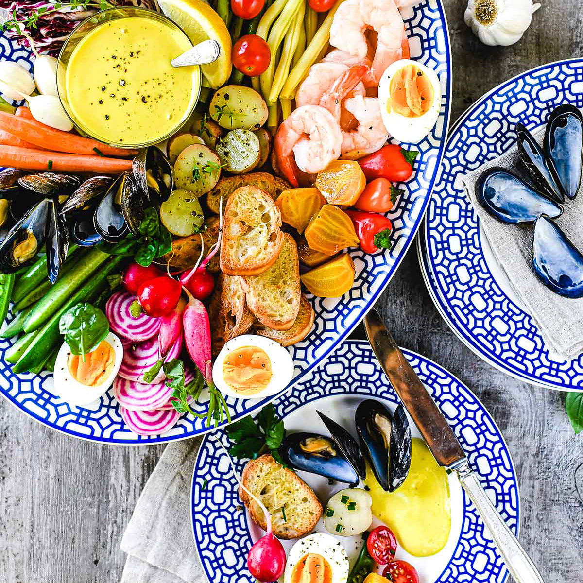 A colorful seafood platter with shrimp, mussels, boiled eggs, vegetables, toasted bread, and a bowl of yellow dipping sauce, served on Newport Garden Gate Rimmed Salad Plates from Caskata Artisanal Home.