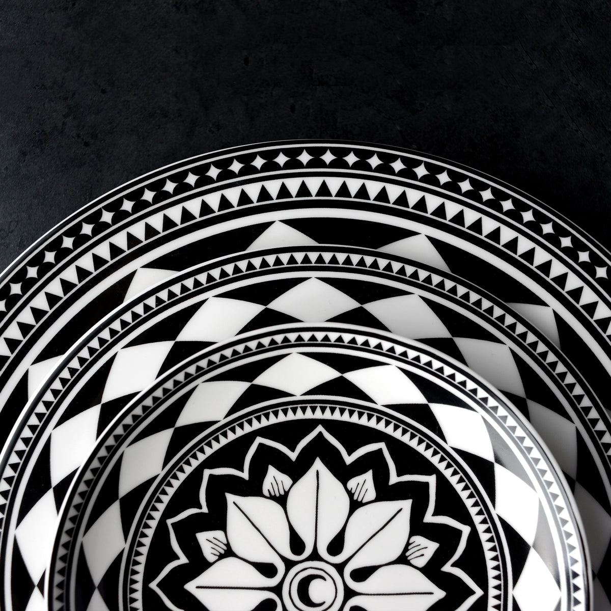 A sophisticated Fez Rimmed Dinner Plate by Caskata Artisanal Home featuring nested black and white plates adorned with intricate geometric and floral Moroccan patterns against a dark background.