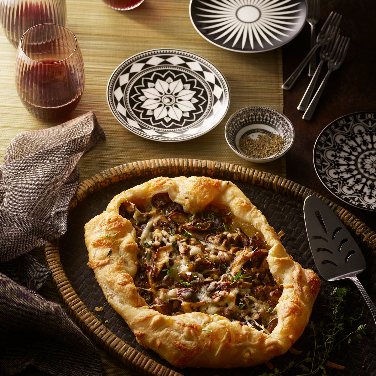 A rustic vegetable galette on a wicker tray, surrounded by Caskata Artisanal Home&#39;s Fez Small Plates featuring bold geometrics, silverware, two glasses of red wine, and a small bowl of herbs on a woven tablecloth.
