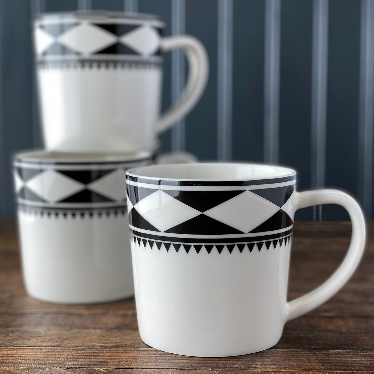 Three white Moroccan-inspired Fez Mugs by Caskata Artisanal Home with black geometric patterns, one in the foreground and two stacked in the background, placed on a wooden surface with a dark striped background. These high-fired porcelain mugs are both dishwasher and microwave safe.