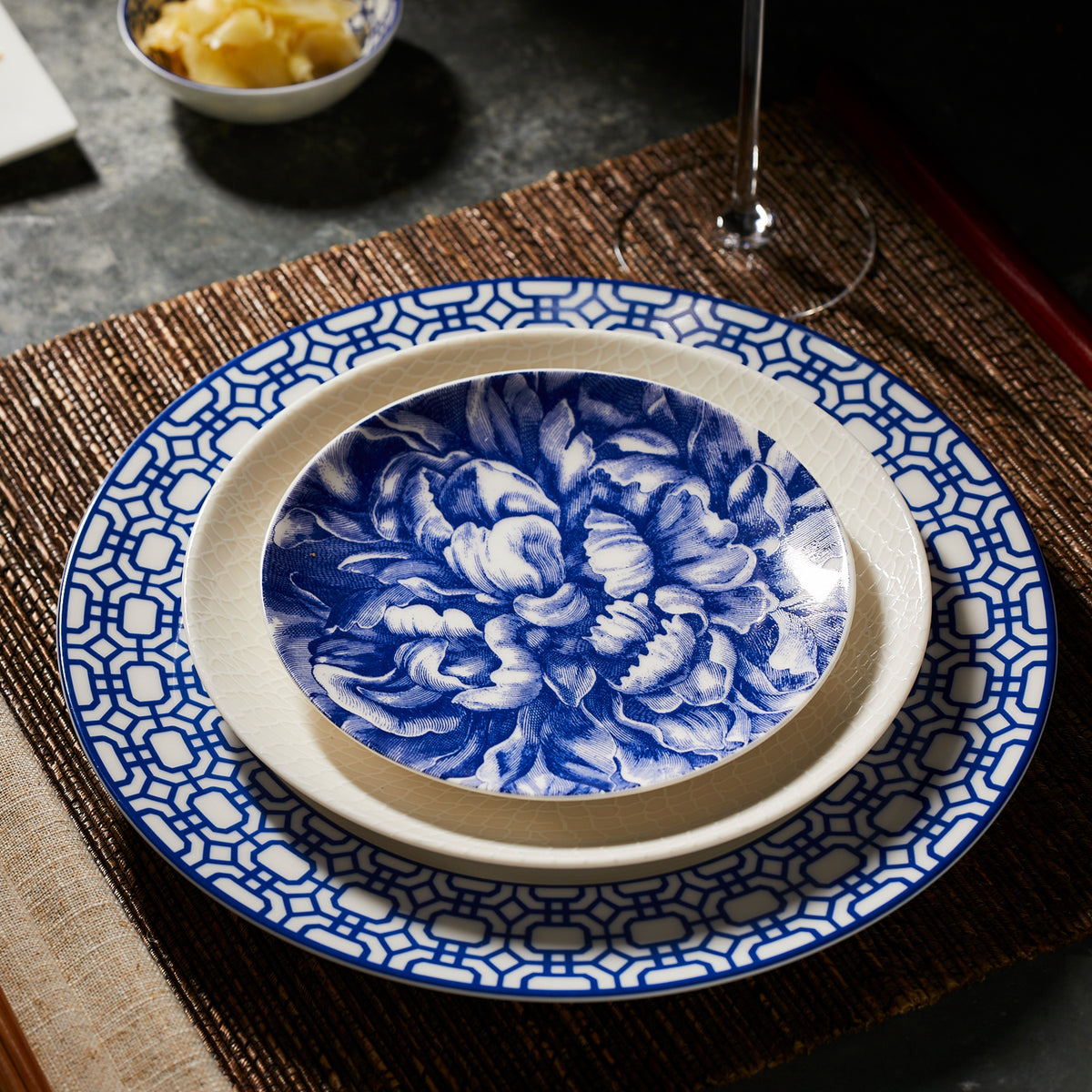 A close-up of a table setting features three nested plates from the coastal collections: a blue and white floral-patterned small bowl, a plain off-white middle plate, and a large Caskata Artisanal Home Newport Garden Gate Rimmed Dinner Plate with blue and white geometric patterns.
