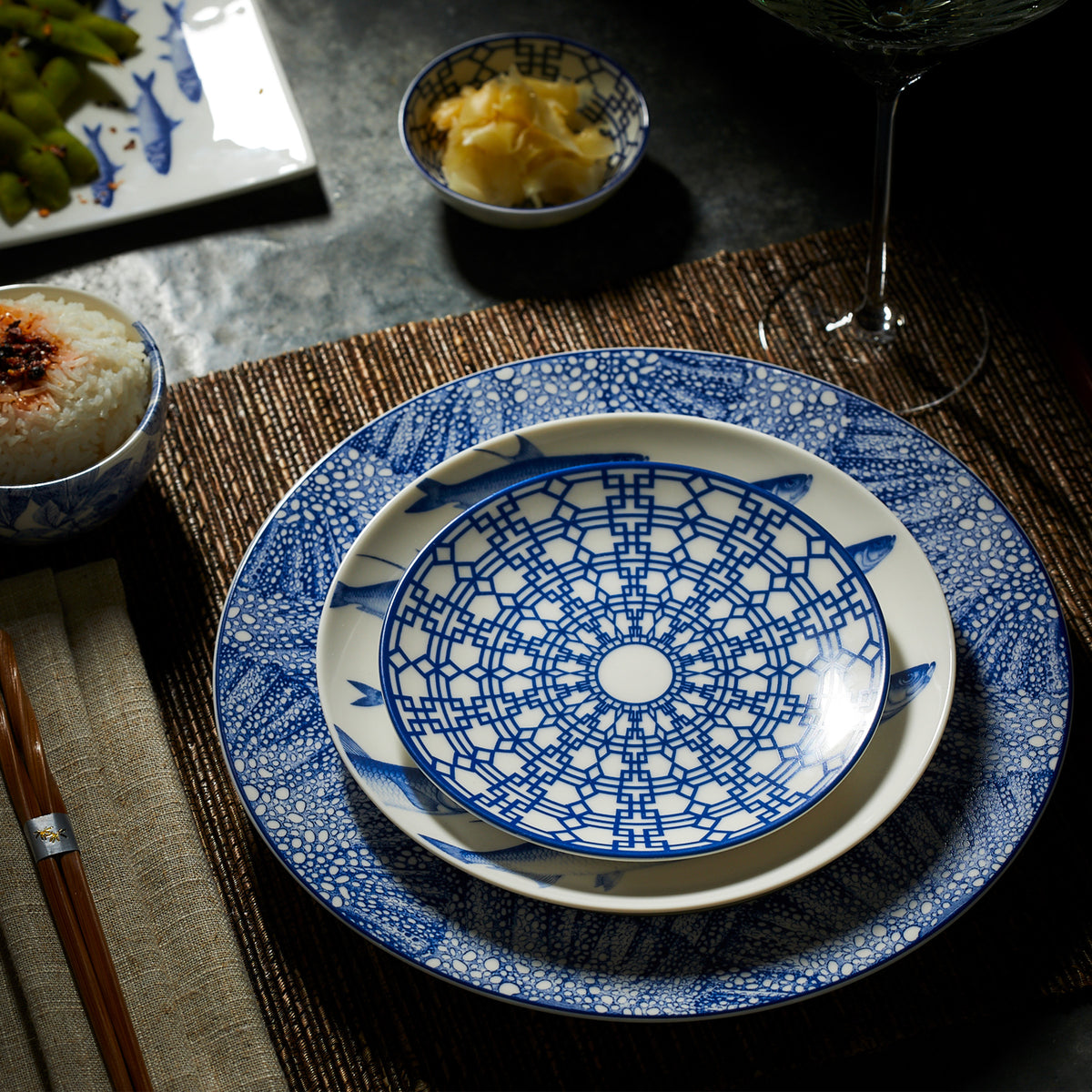 A dining setup featuring intricately patterned blue and white Caskata Artisanal Home Newport Small Plates, reminiscent of the Newport Garden Gate design, placed on a mat. A bowl of rice, a smaller dish with pickles, chopsticks, and part of a glass are visible in the background.