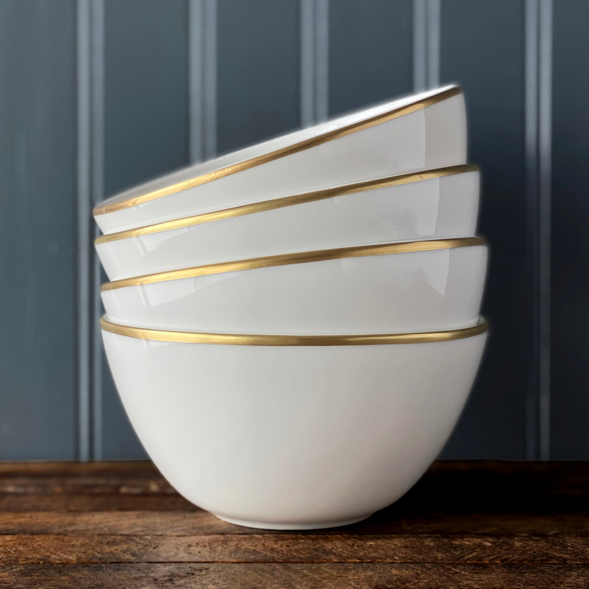 Four stacked white Caskata Artisanal Home Grace Gold Cereal Bowls with gold rims sit on a wooden surface against a dark, vertically paneled background. This sleek, versatile collection is crafted from lead-free high-fired porcelain, ensuring both elegance and durability.