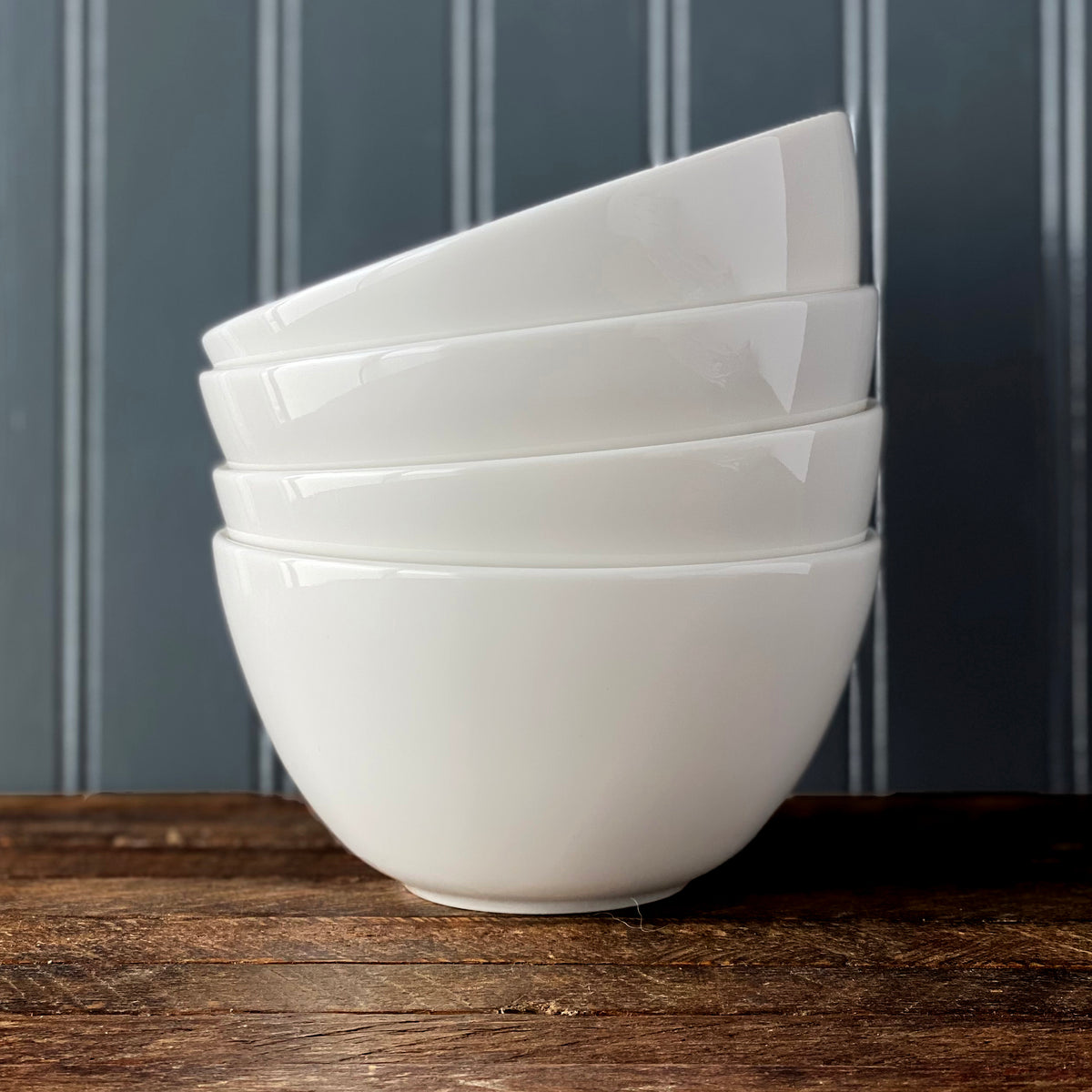 Four sleek, high-fired Grace White Cereal Bowls by Caskata Artisanal Home stacked on a rustic wooden surface with a vertical striped wall background.