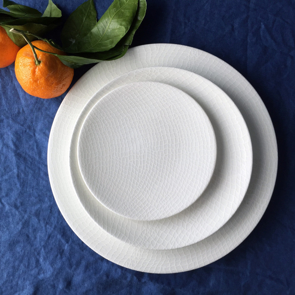 Three white textured plates of varying sizes, crafted from high-fired porcelain, are stacked on a blue tablecloth resembling a fisherman&#39;s net, with two oranges and leaves placed next to them. These plates are known as Catch Small Plates by Caskata Artisanal Home.