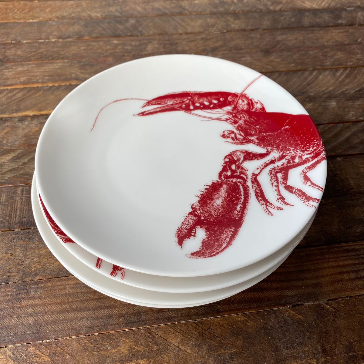 Three white ceramic Lobster Red Small Plates by Caskata Artisanal Home stacked on a wooden surface, each featuring a red lobster design on one side. These heirloom-quality dinnerware pieces evoke a charming seaside style, perfect for any coastal-themed table setting.