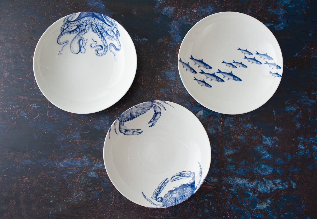 Three white ceramic bowls, accentuated with blue sea life designs, including an exquisite Lucy Entrée Bowl by Caskata Artisanal Home, a school of fish, and crabs, arranged on a dark surface. These pieces are crafted from high-fired porcelain for exceptional durability and elegance.