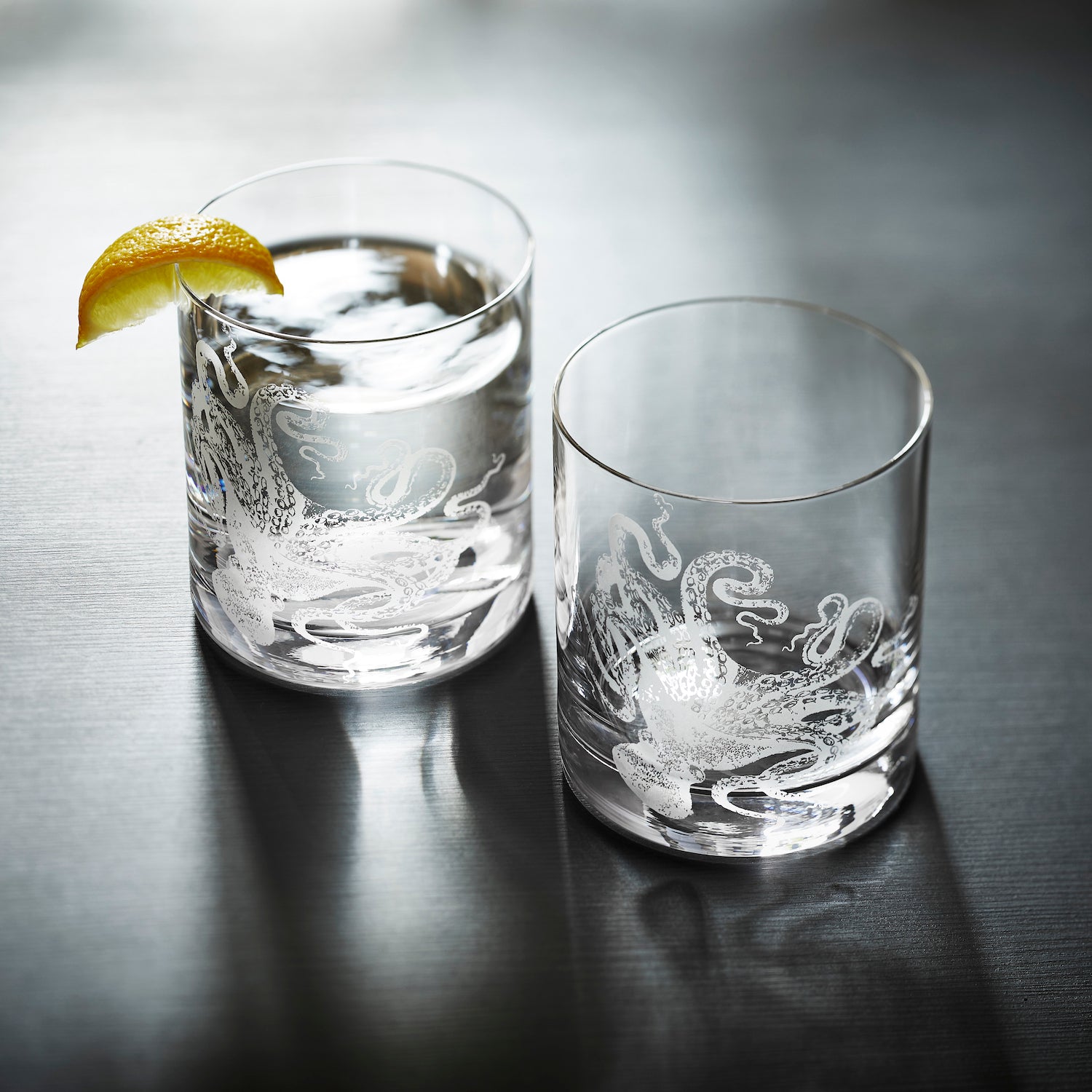 Two Lucy Rocks Glasses, crafted from exquisite crystal glassware by Caskata Artisanal Home, feature intricate octopus designs created using the sand-etching technique. Placed side by side, they merge elegance with a touch of marine mystique.