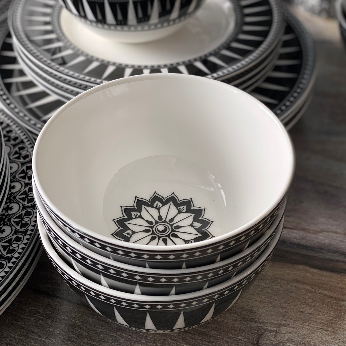 A stack of Marrakech Cereal Bowls by Caskata Artisanal Home, inspired by Art Deco and designed in black and white with geometric patterns. More similarly patterned plates, crafted from high-fired porcelain, are in the background.