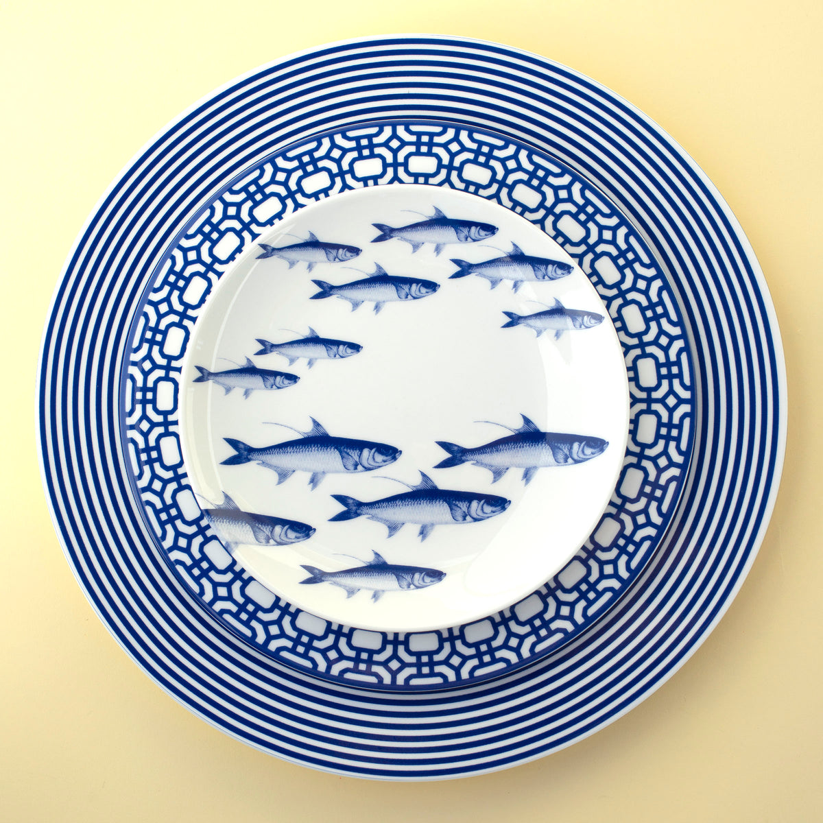 Three nested plates featuring different blue and white patterns; the top plate depicts a school of fish, the middle plate a geometric pattern, and the bottom plate has concentric stripes. The School of Fish Small Plates from Caskata Artisanal Home are microwave safe, heirloom-quality dinnerware that brings both beauty and practicality to any table setting.