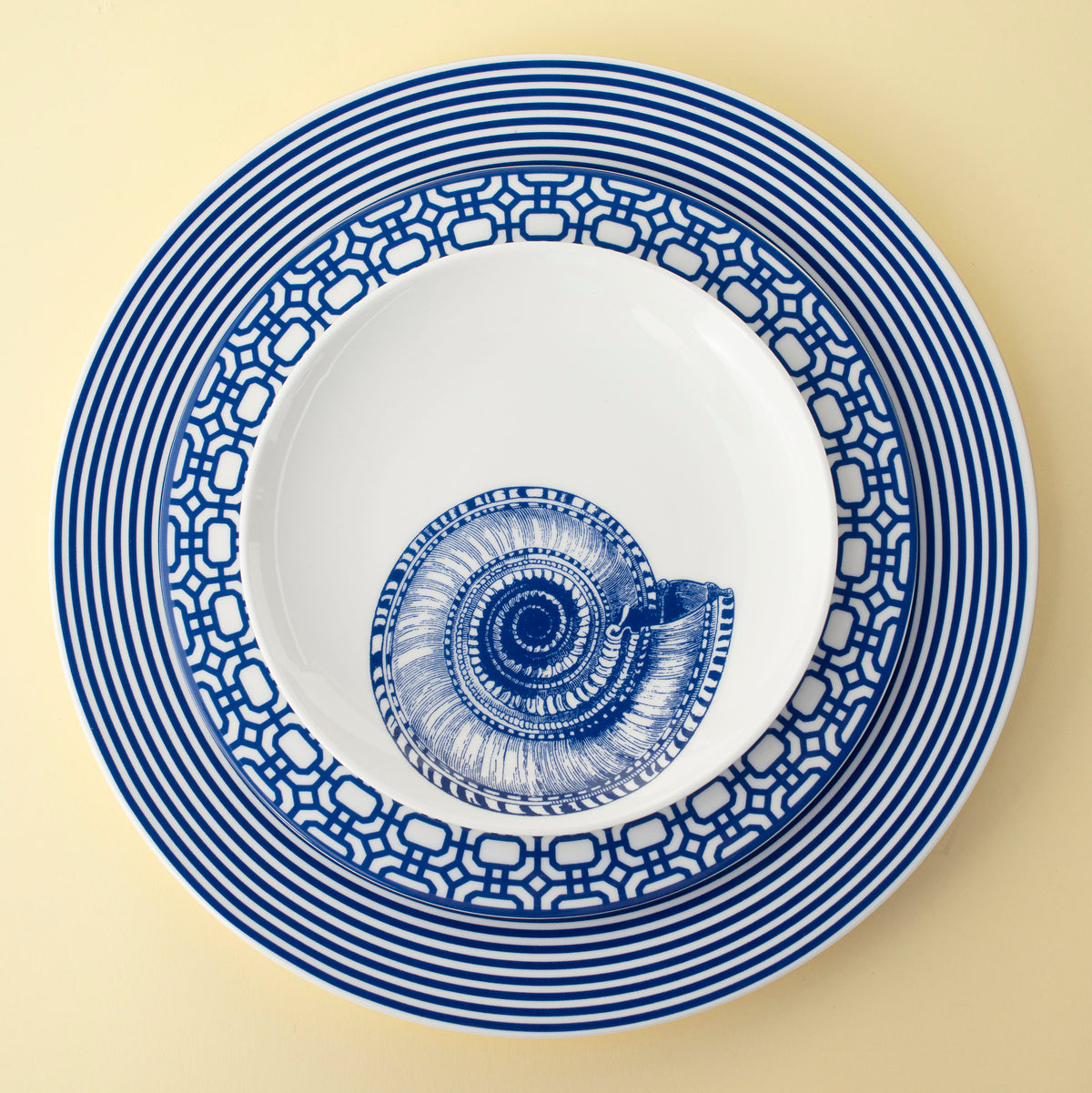 Three blue-and-white plates with geometric and striped patterns are stacked. The smallest plate, part of our beach collection, features a blue nautilus shell design in the center. This Shells Small Plates by Caskata Artisanal Home is set against a light yellow background.