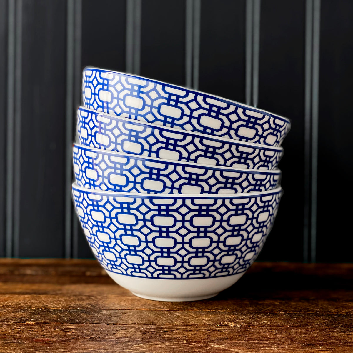 A stack of four blue and white patterned Newport Cereal Bowls by Caskata Artisanal Home, reminiscent of the Newport Garden Gate design, is placed on a wooden surface with a dark, vertically paneled background.