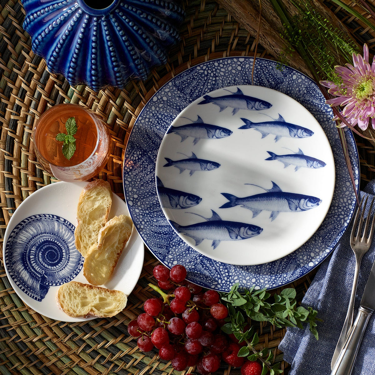 A table setting featuring heirloom-quality dinnerware with blue fish-patterned plates, Shells Small Plates by Caskata Artisanal Home with bread slices, a drink with mint, grapes, a blue cloth napkin, and a purple flower on a woven placemat background.