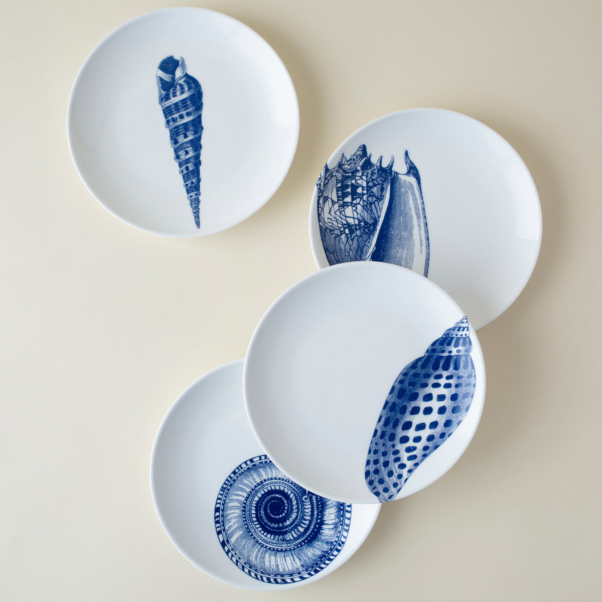Four heirloom-quality Shells Small Plates by Caskata Artisanal Home featuring delicate blue sea shell illustrations are arranged in a loose circular formation on a light-colored surface, making them a stunning addition to your beach collection.