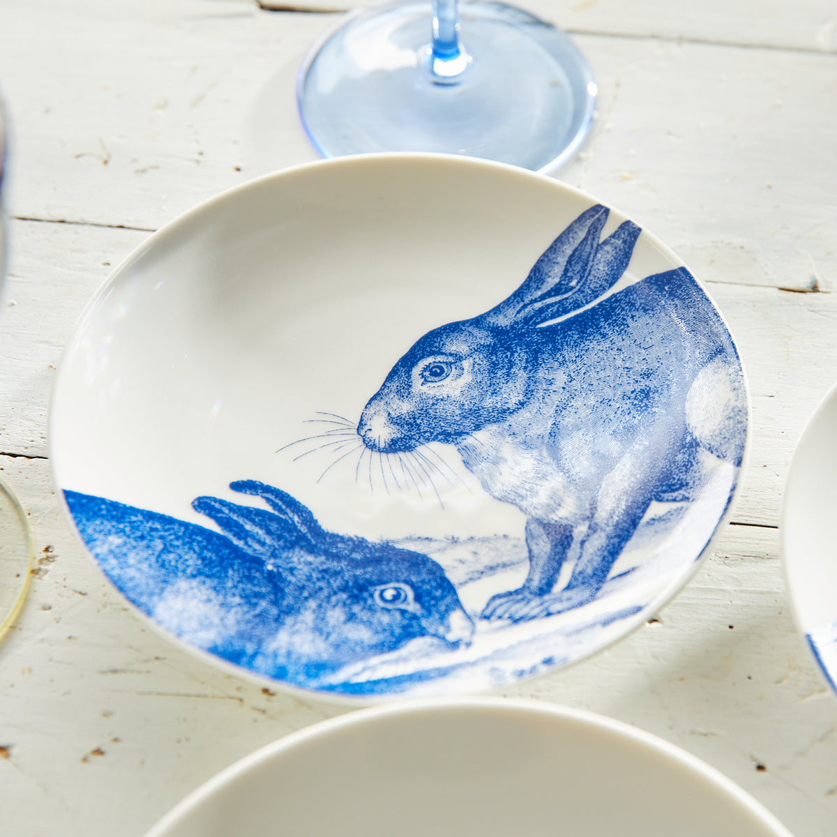 A white bowl with blue illustrations of two rabbits, one sitting upright and the other crouched. The Bunnies Small Plates, crafted from high-fired porcelain by Caskata Artisanal Home, rest on a white wooden surface next to a stemmed glass and other partially visible plates.