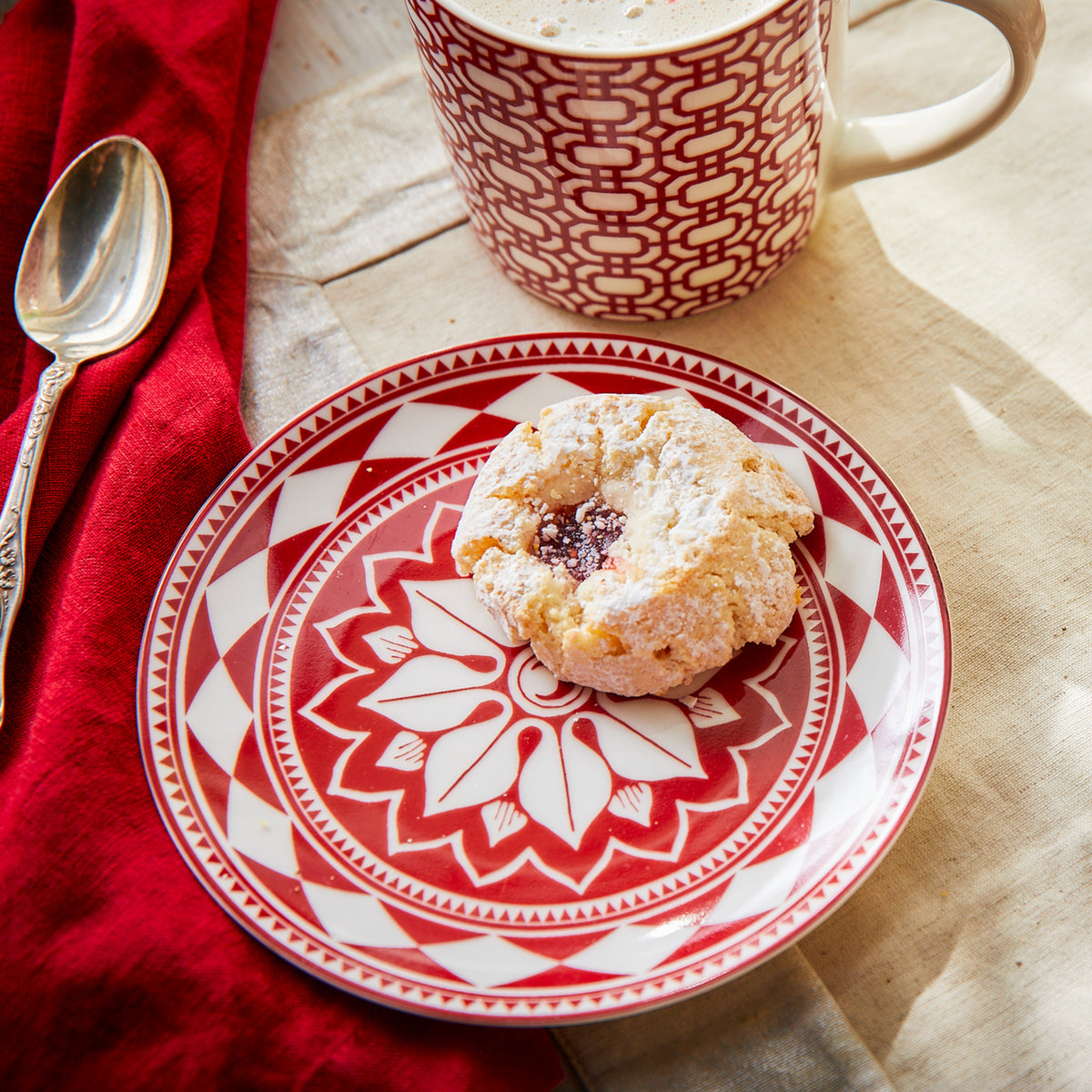 A neatly arranged plate with a single pastry topped with powdered sugar and a dollop of jam sits on a red and white patterned Fez Crimson Small Plate by Caskata Artisanal Home, part of our heirloom-quality dinnerware collection, next to a matching mug and spoon on a tablecloth.