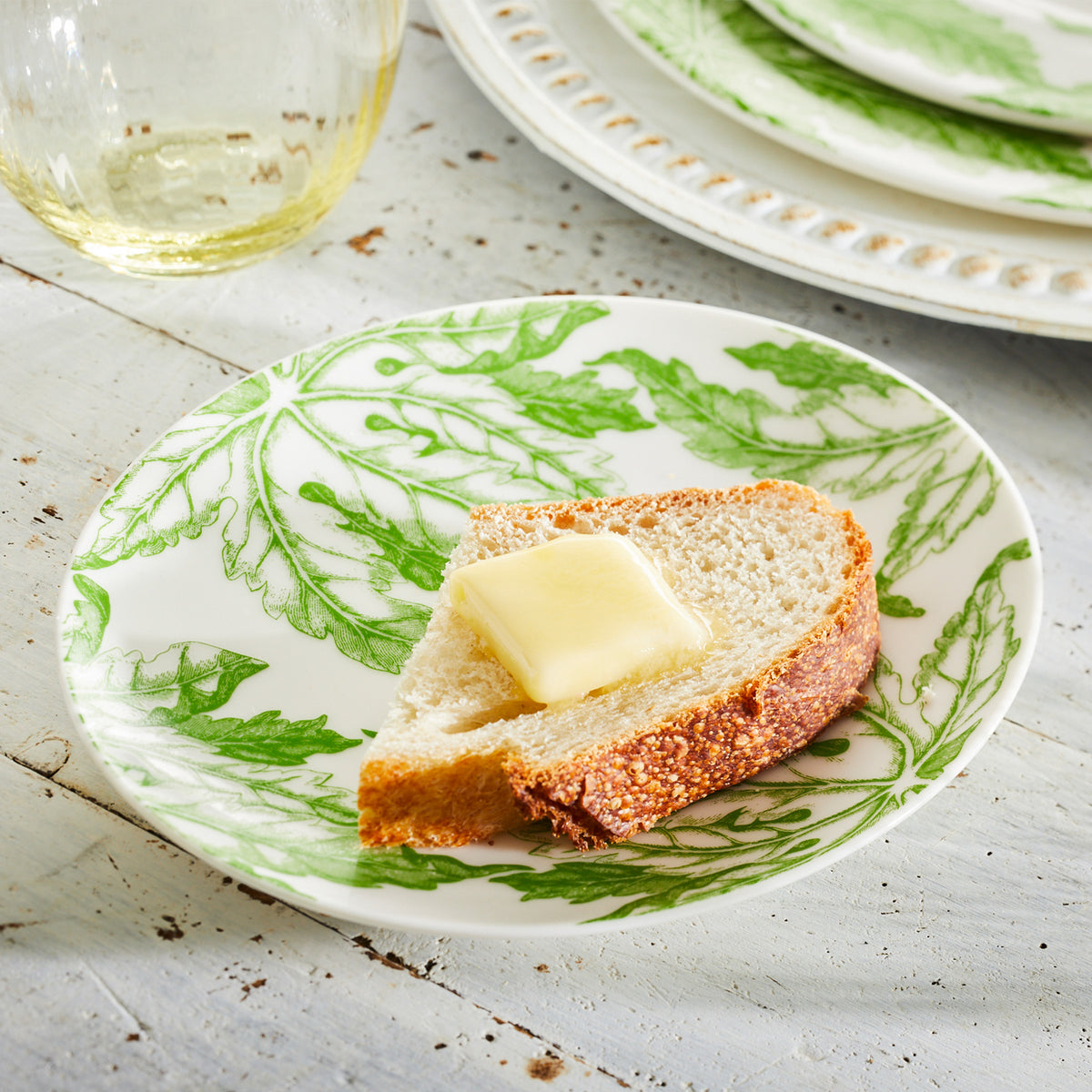 A slice of bread with a pat of butter rests on Freya Small Plates by Caskata Artisanal Home, showcasing a green floral pattern, placed on a white wooden surface. A glass and a plate are partly visible in the background.