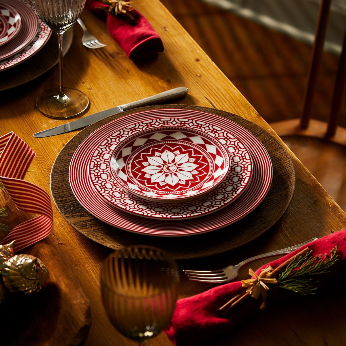 A dining table set with red and white patterned plates, including Caskata Artisanal Home Newport Garden Gate Crimson Rimmed Salad Plates, complemented by red napkins with festive decorations, wine glasses, and silverware.