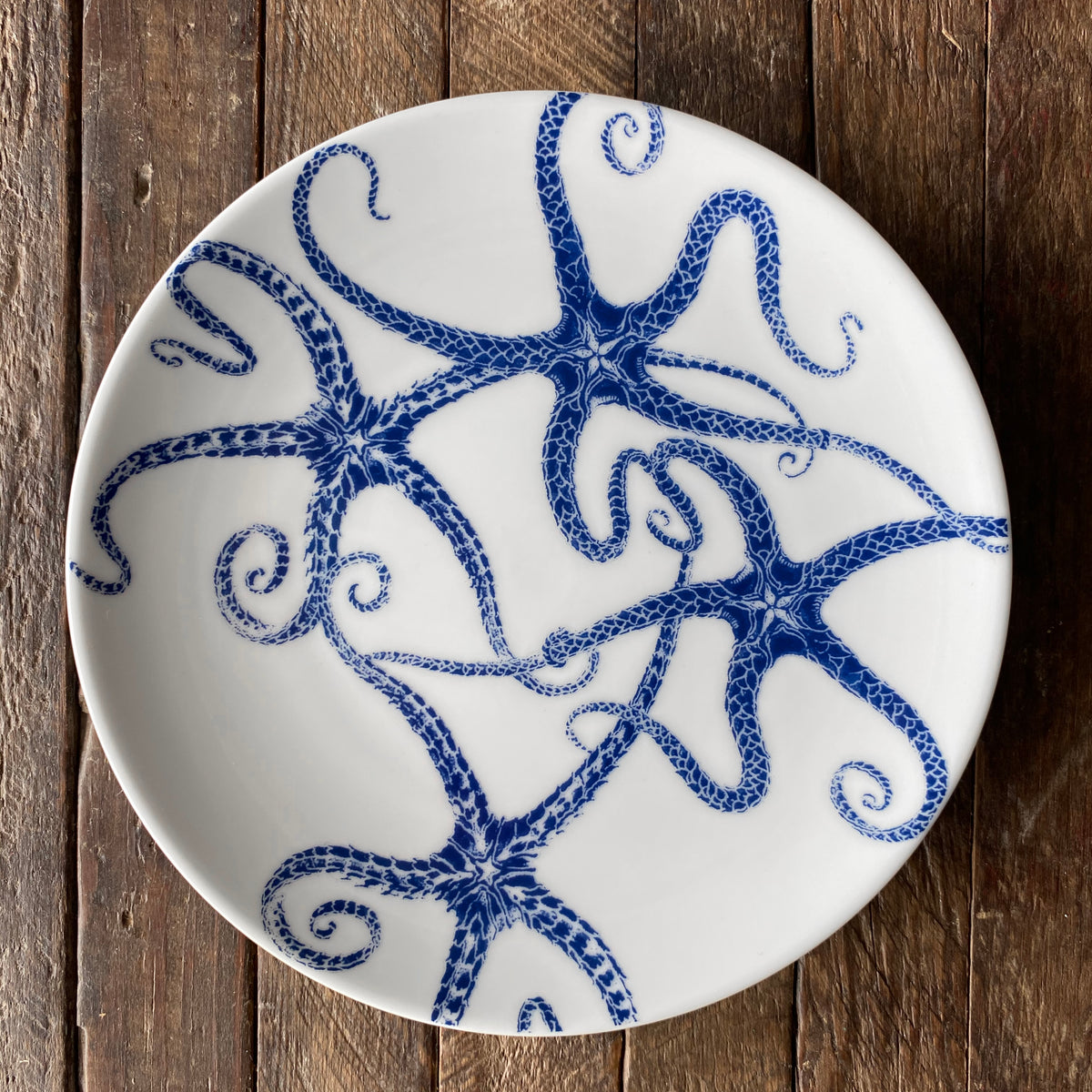 White lead-free porcelain plate with a blue octopus design, placed on a wooden surface. Part of our heirloom-quality dinnerware collection, this piece pairs beautifully with our Caskata Artisanal Home Starfish Small Plates for an ocean-inspired table setting.