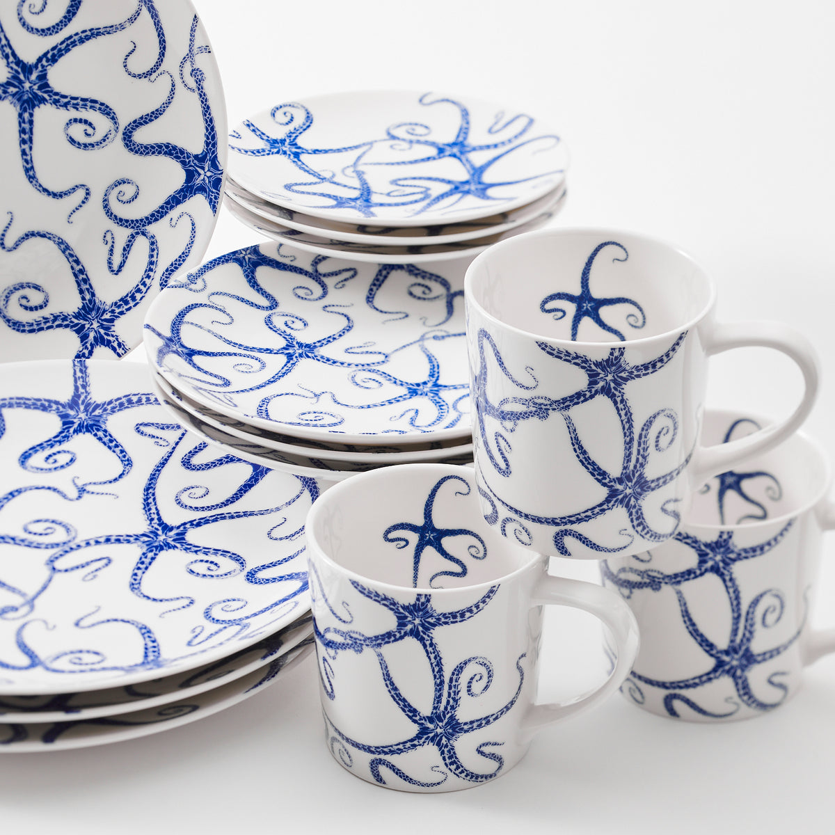 A set of Caskata Artisanal Home Starfish Coupe Dinner Plates and mugs, adorned with blue octopus patterns, arranged in a stack on a white background. Their contemporary shape adds an elegant touch, reminiscent of delicate starfish gracing a coastal tableau.