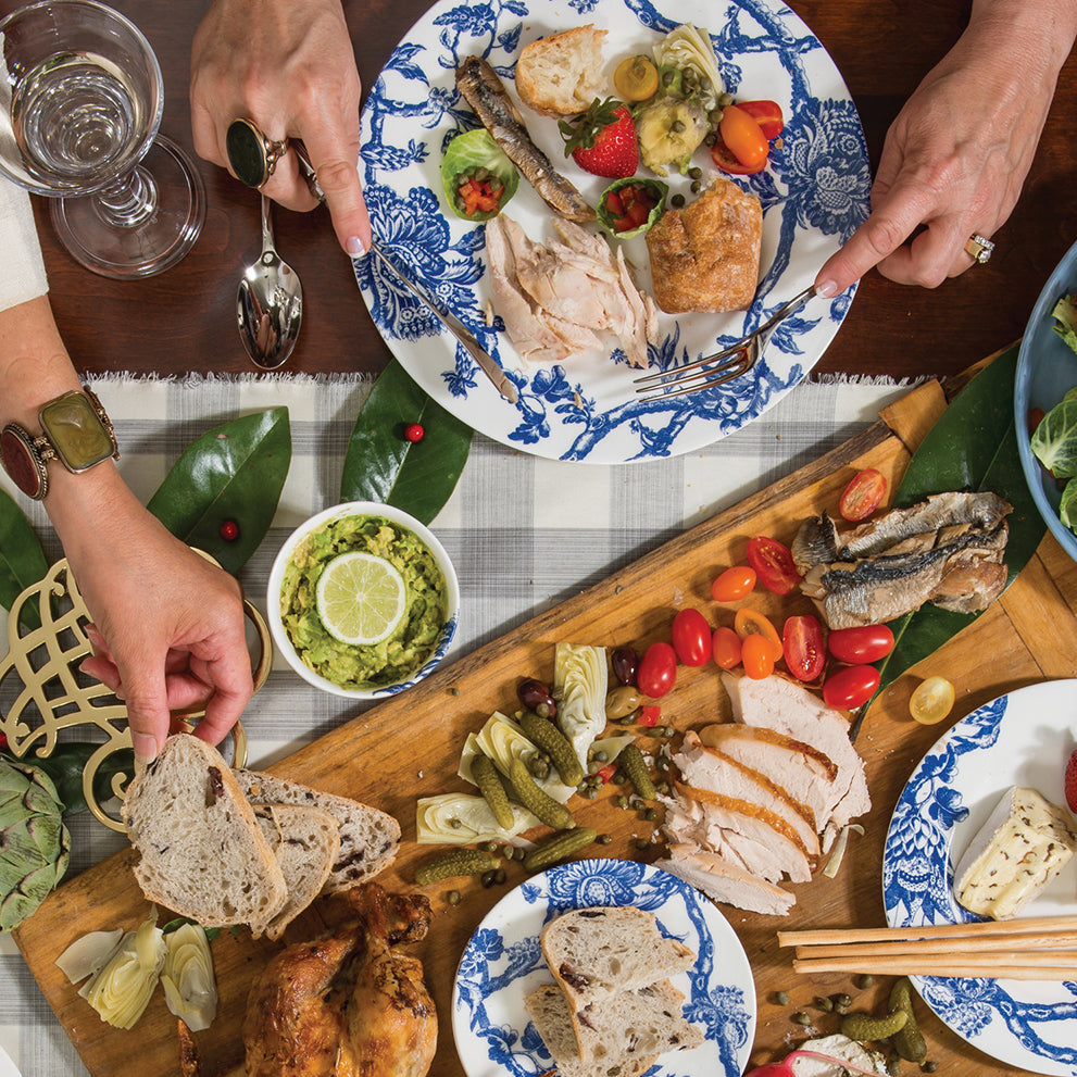 Overhead view of a dining table with premium porcelain dinnerware showcasing plates of food being shared. Dishes include bread, sliced meat, salad, tomatoes, pickles, and other garnished foods. Three hands holding utensils are visible, featuring Arcadia Small Plates by Caskata Artisanal Home.