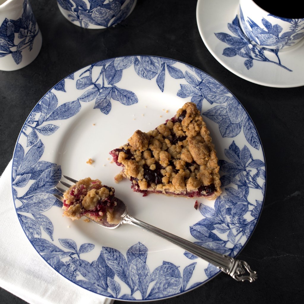 A slice of berry crumble pie on an Arbor Rimmed Salad Plate by Caskata Artisanal Home, accompanied by a fork with a bite-sized portion, sits on a dark surface next to a cup of coffee. This premium porcelain piece adds an heirloom feel to the delightful scene.