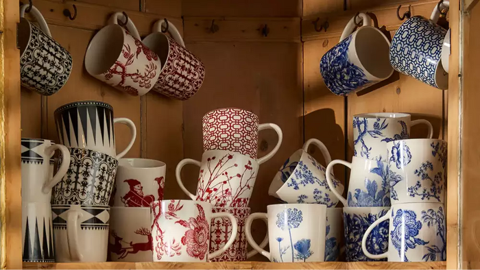 A wooden cabinet filled with Caskata oversized porcelain mugs in bold red, blue, and black patterns