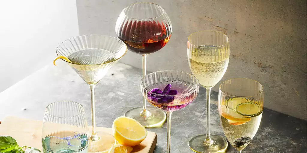 A variety of cocktails served in elegant glassware, including martini, coupe, and wine glasses, styled with citrus, herbs, and floral garnishes on a sunlit table