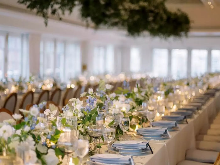 Elegant long dining table decorated with white and blue flowers, candles, and overhead greenery