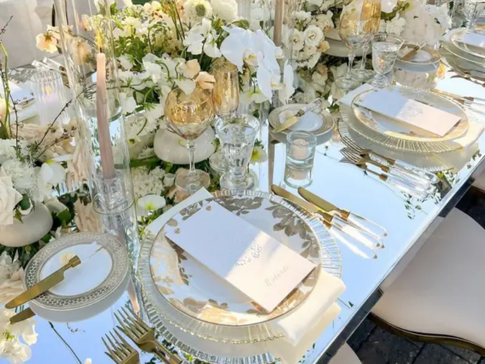 Elegant outdoor wedding table setting with gold flatware, mirrored table, white florals, and crystal glassware in natural sunlight