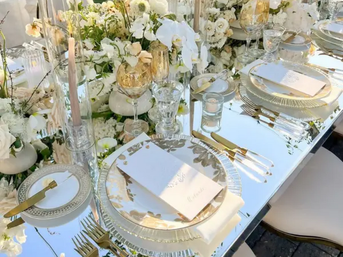 Elegant outdoor wedding table setting with gold flatware, mirrored table, white florals, and crystal glassware in natural sunlight