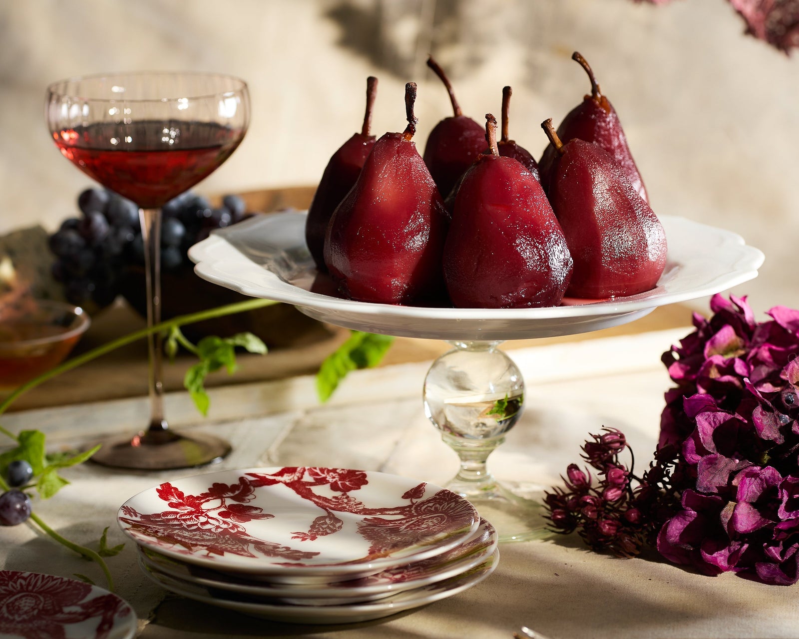 Red wine-poached pears on a glass pedestal cake stand, next to a glass of red wine and a stack of patterned plates.