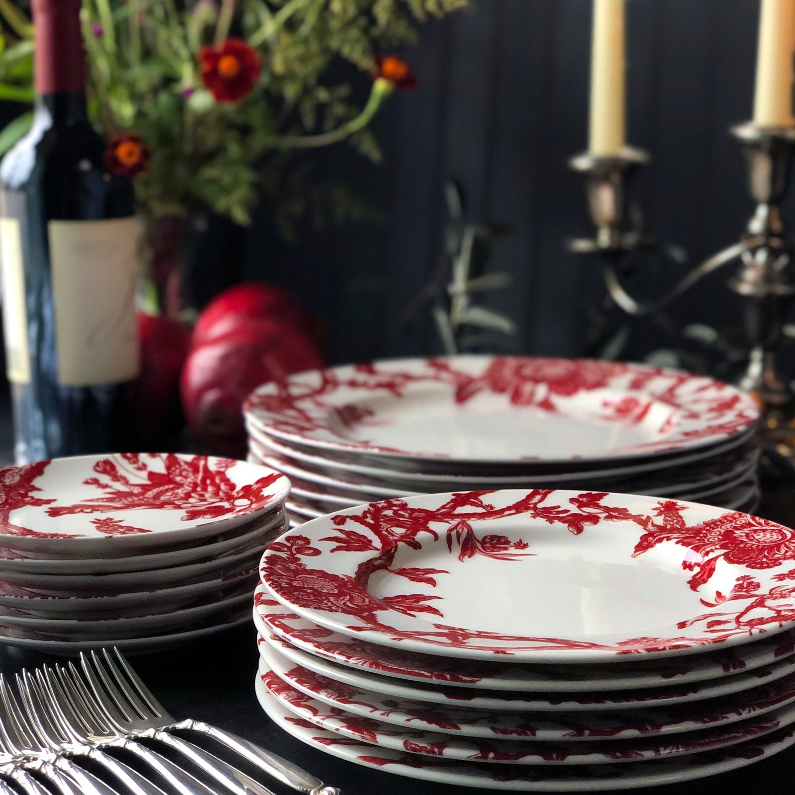 Stacks of decorative white plates with red floral patterns, placed near a bundle of silver forks and candles in a dining setting, with a wine bottle and floral arrangement in the background.