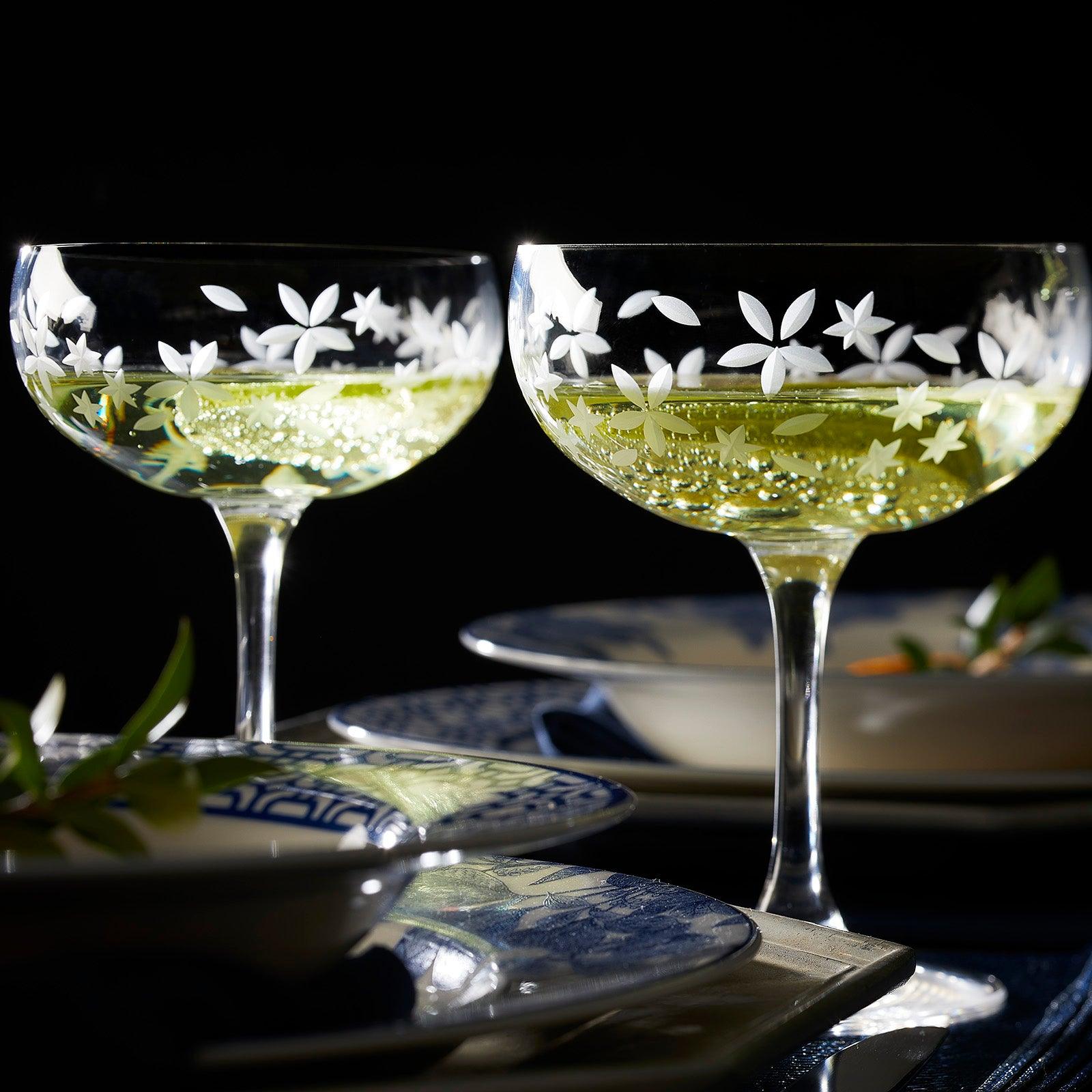 Two floral-etched champagne glasses filled with bubbly liquid are placed on a table set with plates, set against a dark background.