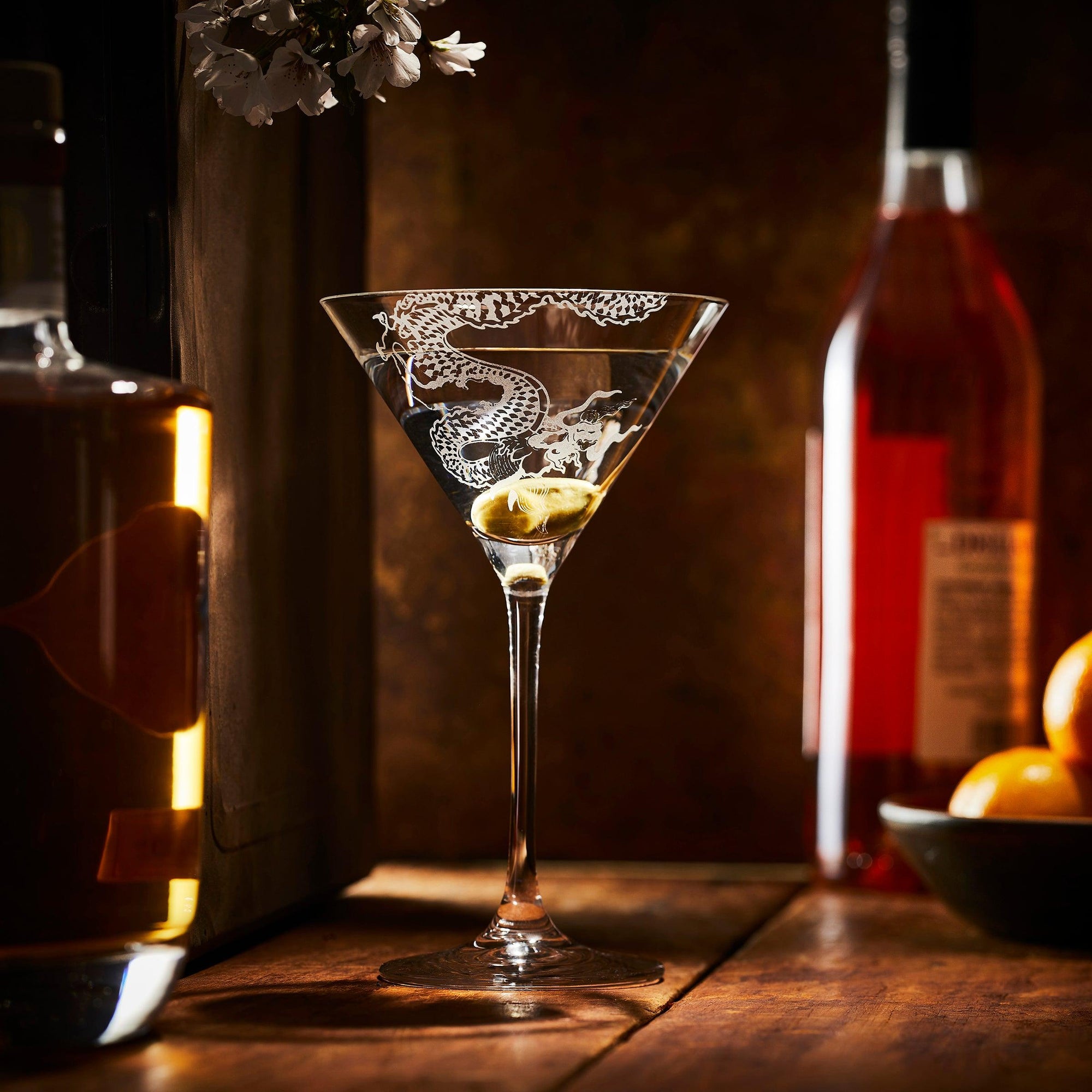 Elegant martini glass with a dragon design, containing a cocktail, on a bar top with bottles and oranges in the background.