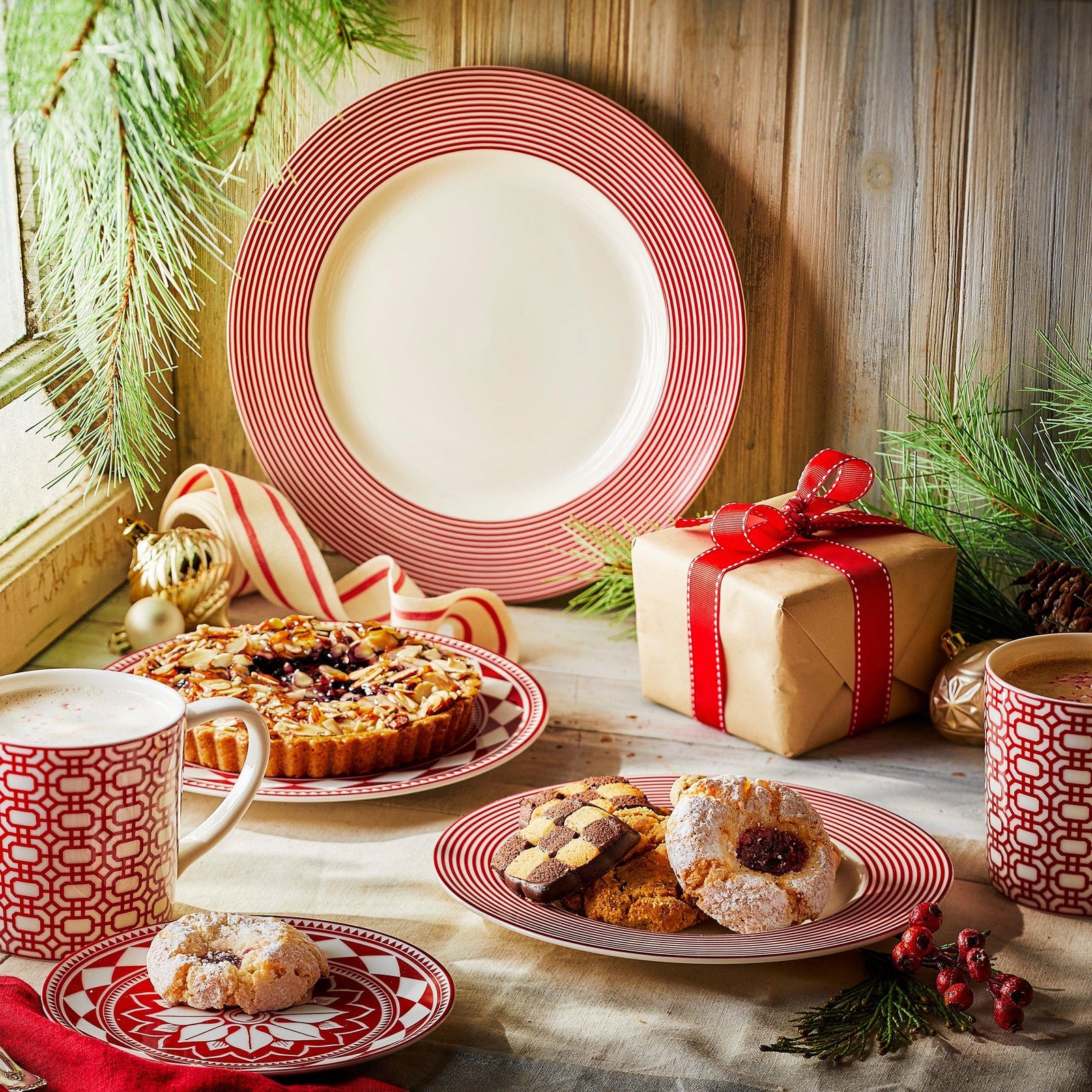 Festive holiday table with red and white plates, various cookies, a tart, two mugs of beverages, a wrapped gift, and pine branch decorations.