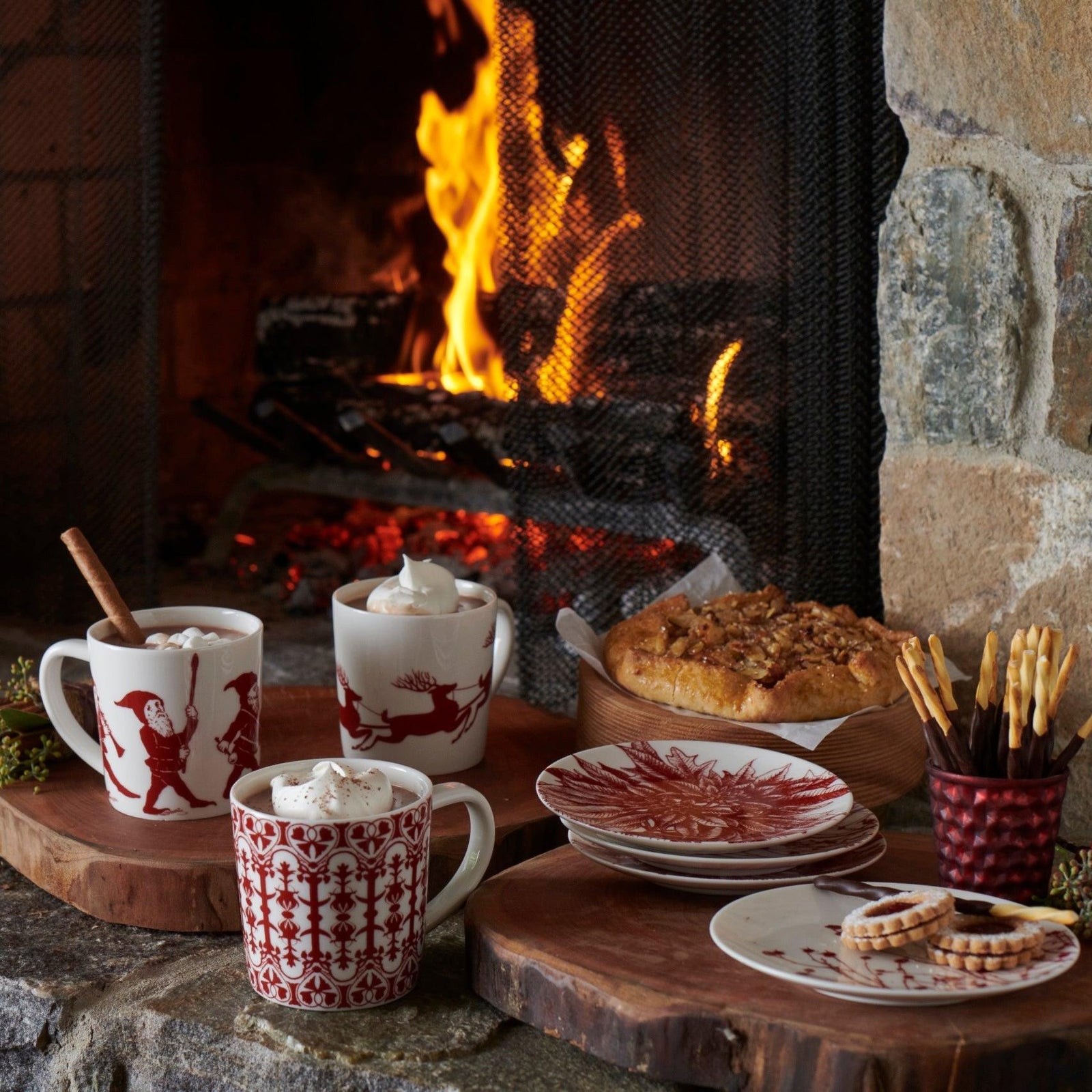 A cozy fireplace scene with mugs of hot chocolate, a cake, cookies, and plates on a stone hearth.