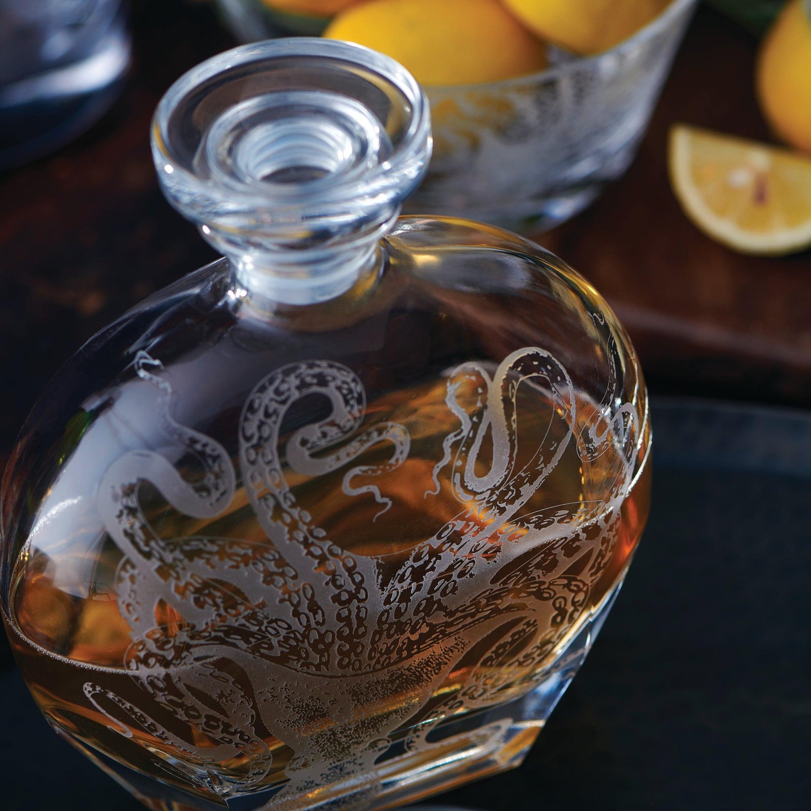 A glass decanter filled with brown liquid, featuring an etched design of an octopus. A bowl of lemons and a lemon wedge are visible in the background.