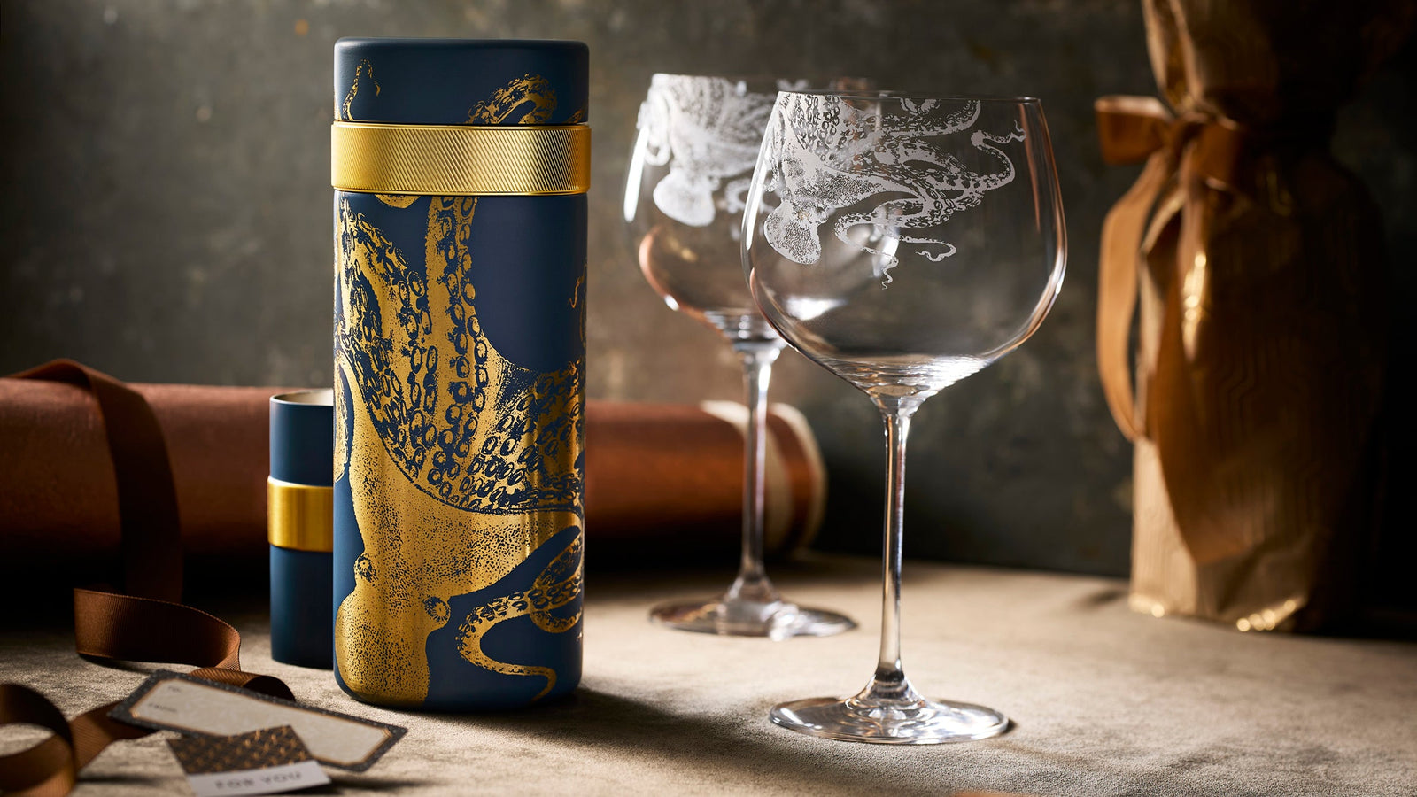 A blue and gold cylinder with an octopus design sits beside two etched wine glasses and wrapped bottles on a beige surface under soft lighting.