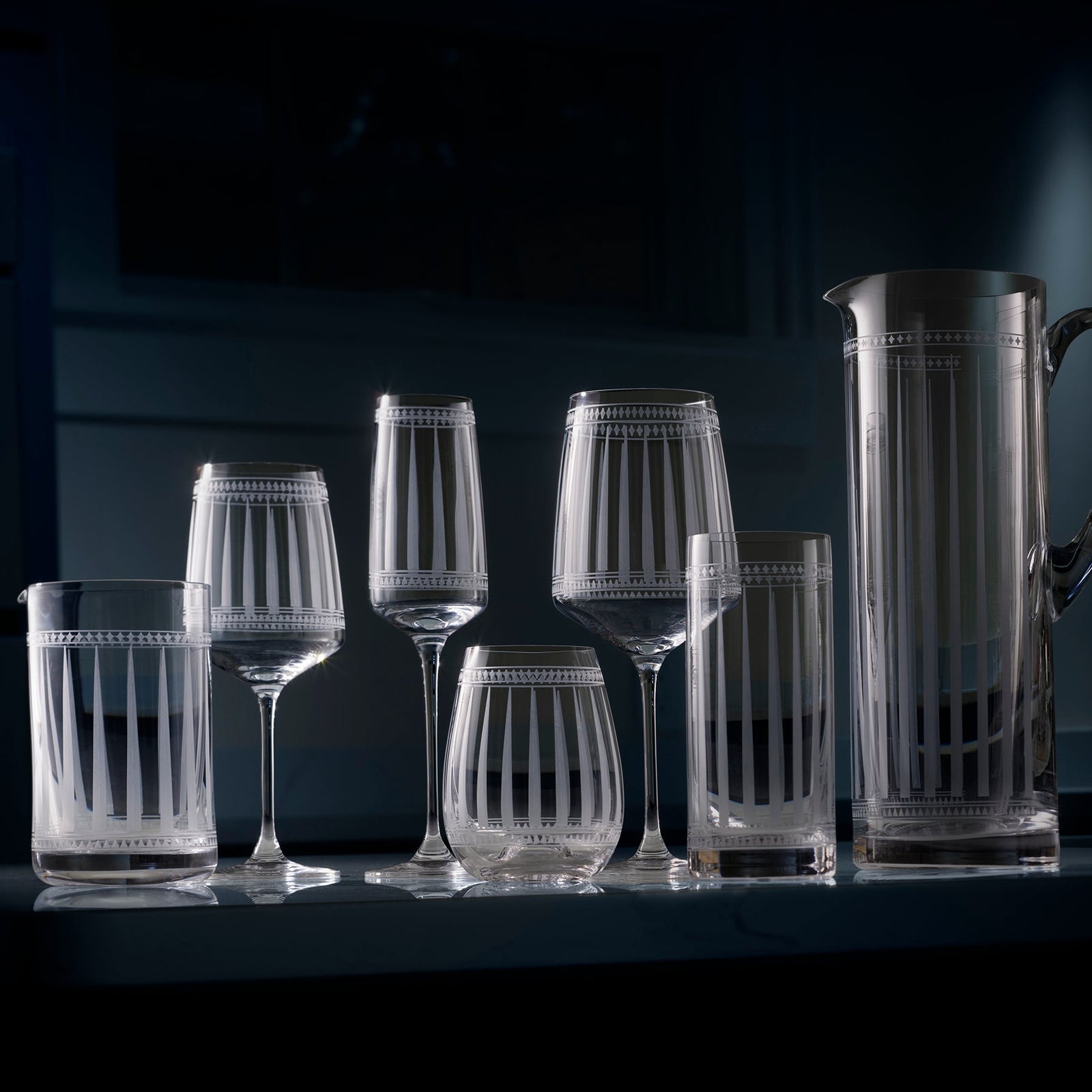 A set of etched glassware, including six glasses and a pitcher, arranged on a dimly lit surface.
