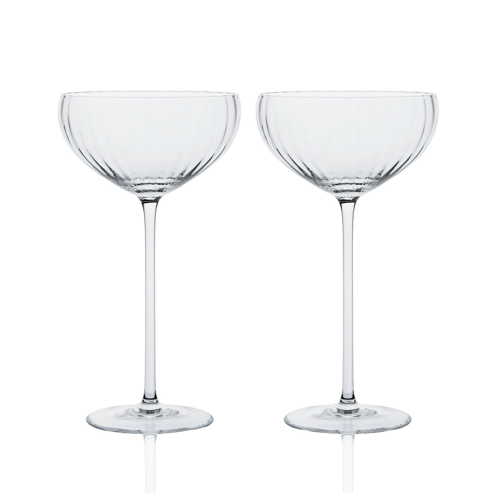 Two empty, clear crystal coupe glasses with ribbed detailing, standing side by side on a reflective surface.