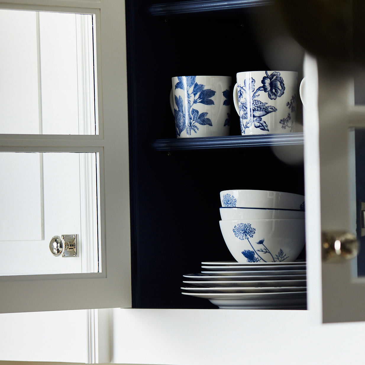 Open kitchen cabinet displaying neatly stacked white dishes and bowls, along with mugs decorated with blue floral patterns.