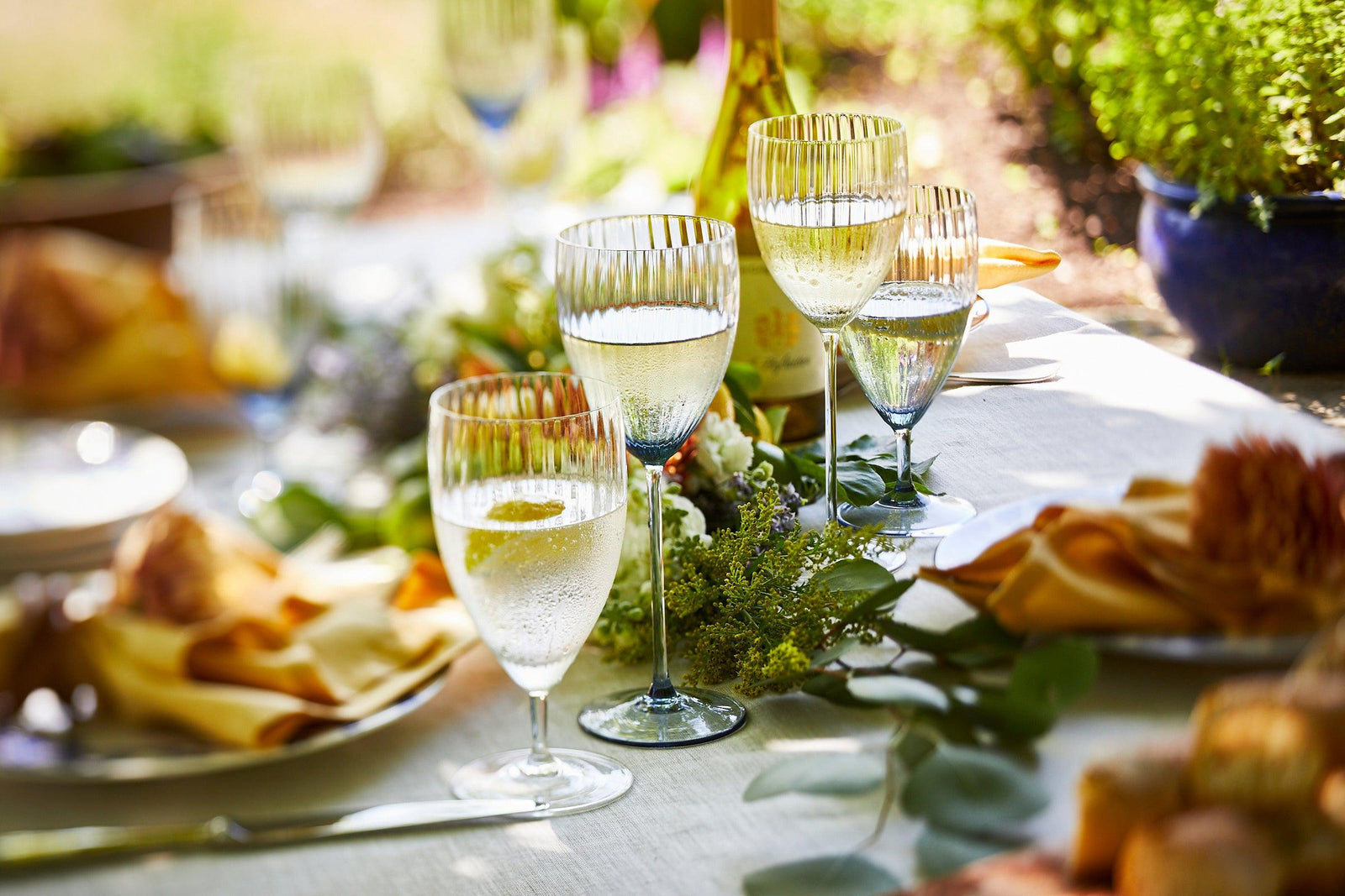 A sunlit table setting with filled wine glasses, a wine bottle, plates, yellow napkins, and floral decorations outdoors.