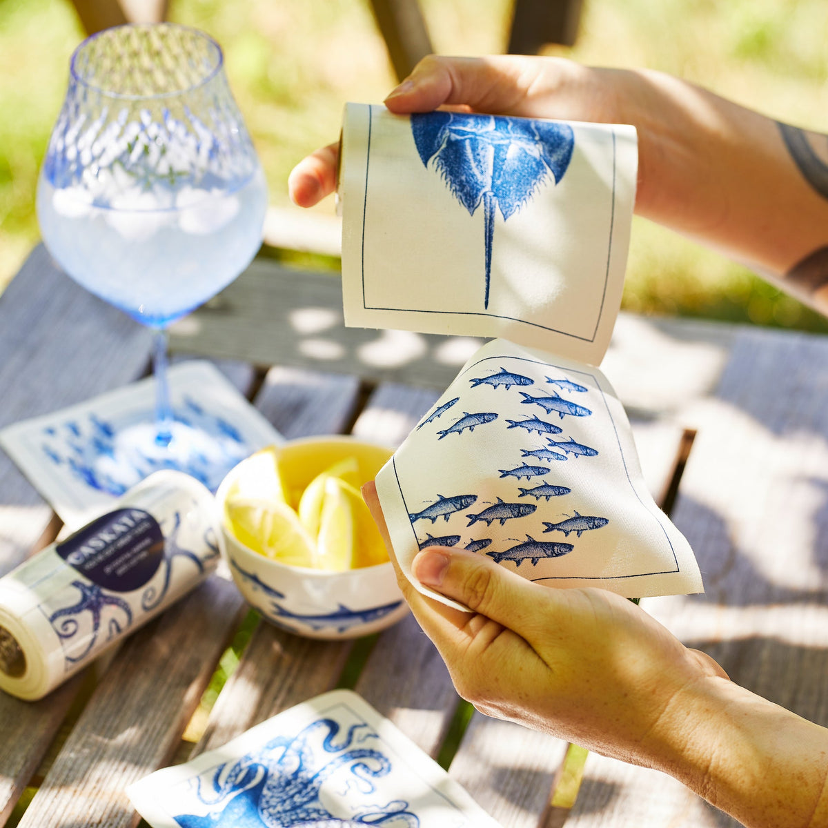 A person is holding a blue and white Coastal Cocktail Napkin Roll by MY DRAP on a table with coastal patterns.