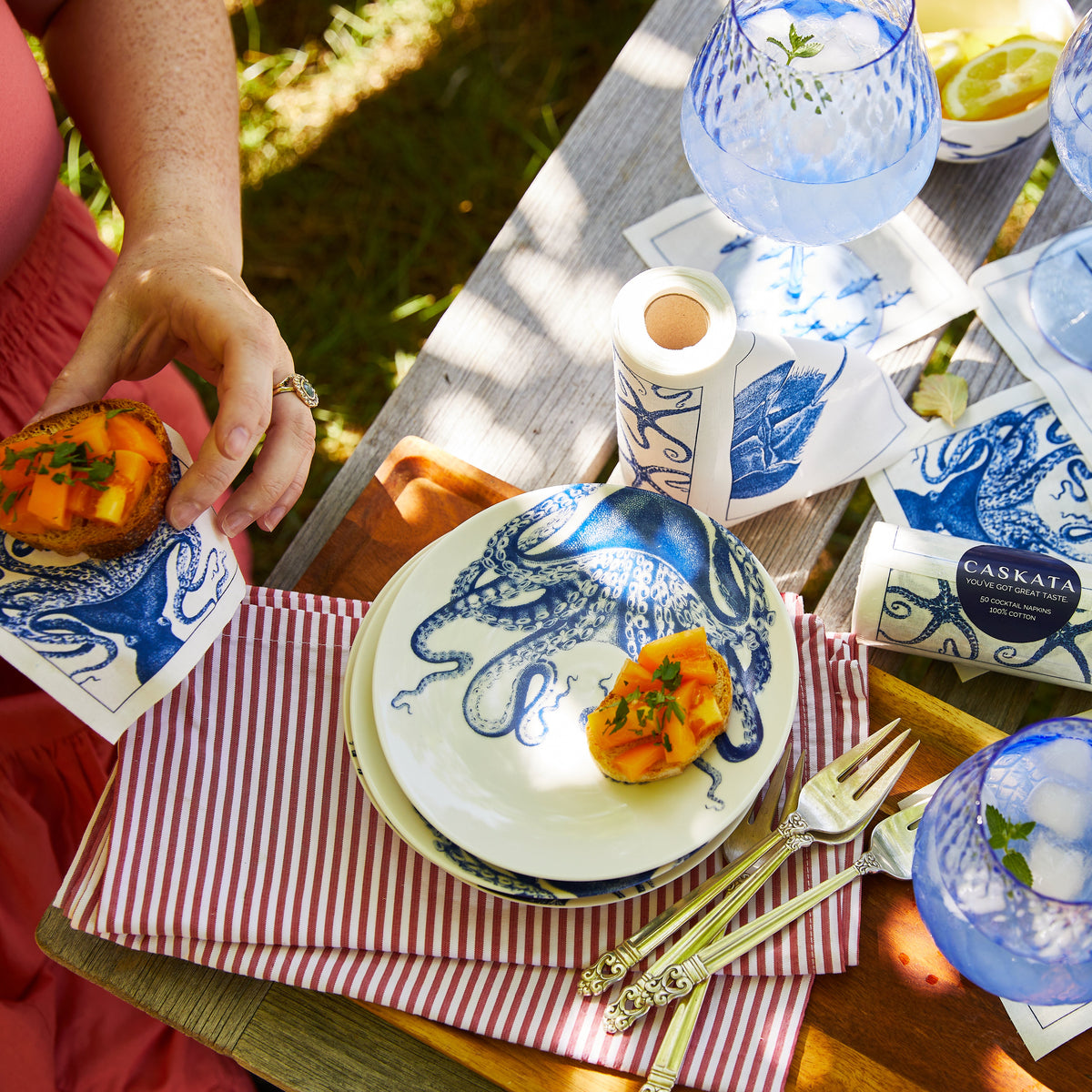 A woman hosting a dinner party, holding a plate of food with MY DRAP Coastal Cocktail Napkin Roll.