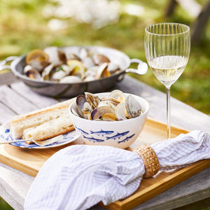 A tray holding a School of Fish Cereal Bowl by Caskata filled with clams, a plate with bread slices, a cloth napkin, and a glass of white wine sits on an outdoor table, with a pan of clams in the background. The setting showcases an elegant dinnerware collection perfect for alfresco dining.