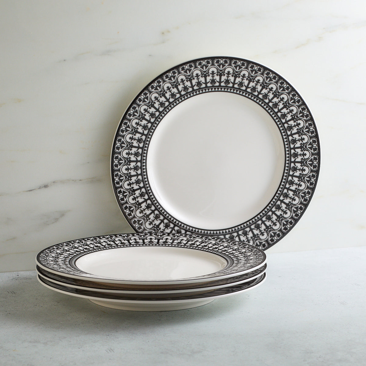 Four Casablanca Rimmed Salad Plates made of lead-free porcelain, with intricate black floral designs on the rims, stacked with one plate resting upright against a white marble backdrop. These pieces feature exquisite hand decorated details from Caskata Artisanal Home.